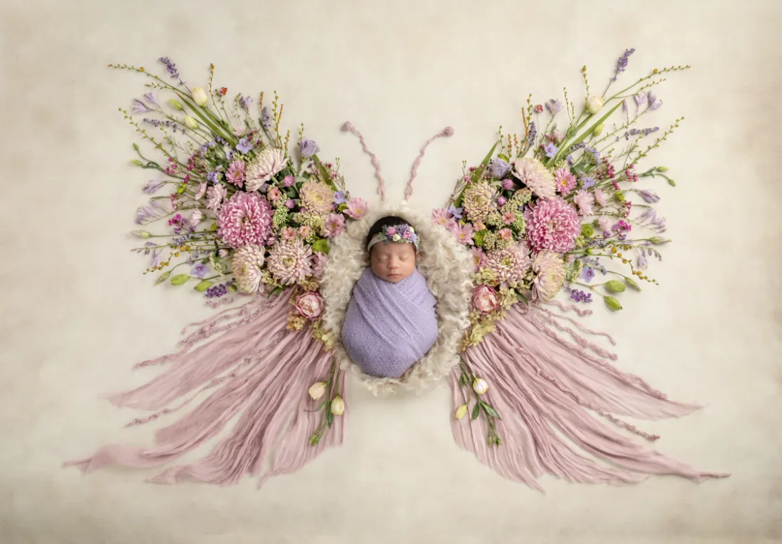 Newborn baby in purple swaddle, surrounded by beautiful spring flowers in the shape of a butterfly